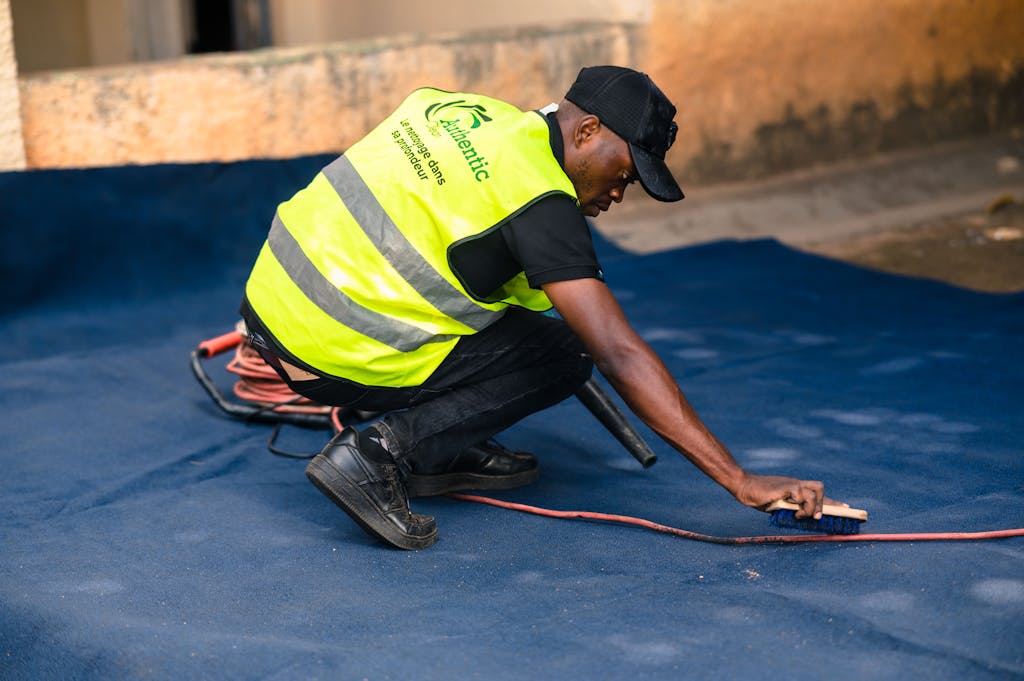 A worker dressed in a safety vest cleans a blue carpet outdoors with a brush.
